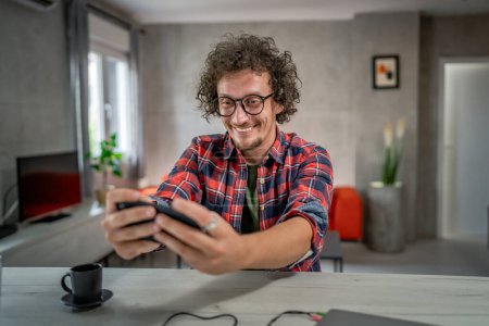 Téléchargez les photos : Homme caucasien assis à la maison jouer à des jeux vidéo sur smartphone téléphone mobile - en image libre de droit