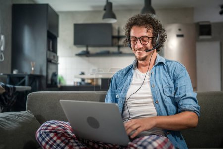 Photo for One man caucasian male freelancer work from home with headphones on his head on laptop computer customer support dispatcher while sit on sofa bed at home in shirt and pajamas happy smile confident - Royalty Free Image