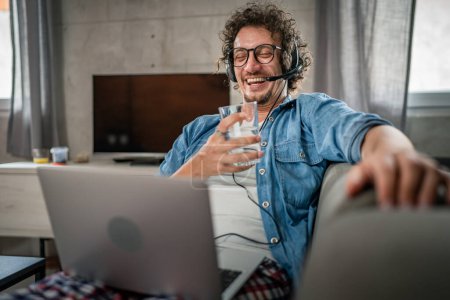 Photo for One man caucasian male freelancer work from home with headphones on his head on laptop computer customer support dispatcher while sit on sofa bed at home in shirt and pajamas happy smile confident - Royalty Free Image