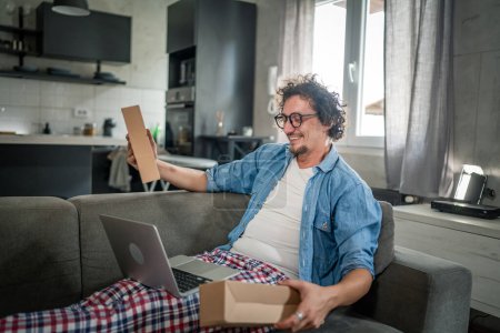 Photo for One caucasian male adult man sit at home on the sofa bed receiving package by post product he ordered online or special gift happy smile real people delivery service concept - Royalty Free Image