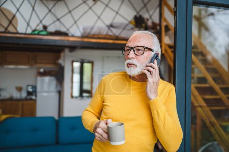 Photo for One man senior caucasian male with gray beard stand on balcony at door in day use mobile smart phone to make a call talk copy space hold cup of coffee worried thoughtful - Royalty Free Image