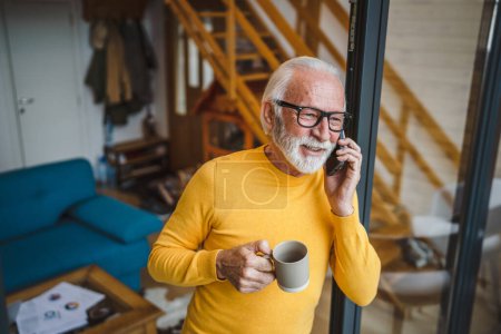 Téléchargez les photos : Un homme homme caucasien senior avec barbe grise debout sur le balcon à la porte dans la journée sourire heureux utiliser téléphone intelligent mobile pour faire un appel parler copie espace tenir tasse de café matin routine quotidienne - en image libre de droit