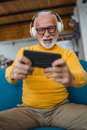 Téléchargez les photos : Un vieil homme caucasien âgé assis à la maison sourire heureux jouer à des jeux vidéo loisirs s'amuser tenir téléphone mobile smartphone avoir un espace de copie amusant - en image libre de droit