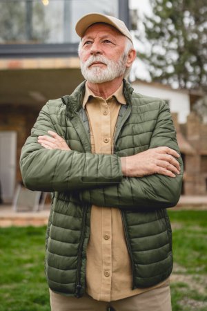 Téléchargez les photos : Un homme caucasien supérieur moderne portrait de grand-père masculin avec barbe et chapeau cap stand en face de sa maison dans la veste de jour heureux confiant copie espace bras croisés - en image libre de droit