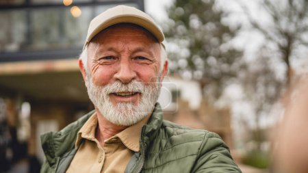 Photo for One modern senior caucasian man portrait of male grandfather with beard and hat cap stand in front of his house in day wear jacket happy confident copy space - Royalty Free Image