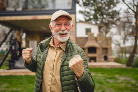 Téléchargez les photos : Un homme caucasien senior moderne portrait de grand-père masculin avec barbe et chapeau se tenir devant sa maison en veste de jour heureux espace de copie confiant célébrer victoire - en image libre de droit