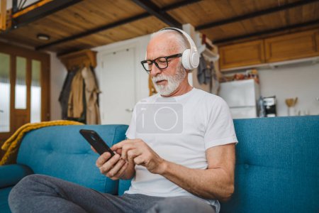 Photo for One senior man with headphones use smsrtphone to play music at home - Royalty Free Image