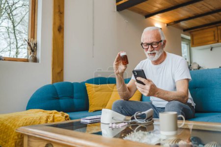 Photo for One senior man sitting at home on sofa bed holding bottle of medicine pills medicament or supplement and smartphone read label instructions of drug tablet painkillers or vitamin ready to take medicine - Royalty Free Image