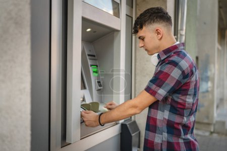Téléchargez les photos : Homme adolescent étudiant en utilisant la carte de crédit et retirer de l'argent au guichet automatique - en image libre de droit