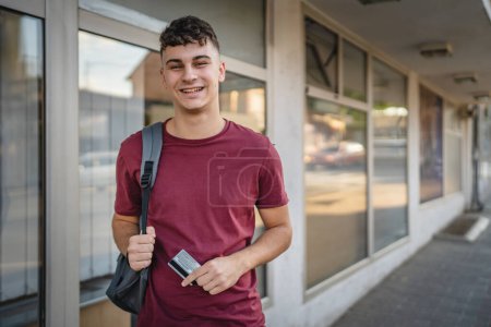 Téléchargez les photos : Portrait de l'homme adolescent debout devant la banque ATM détenir carte de crédit - en image libre de droit