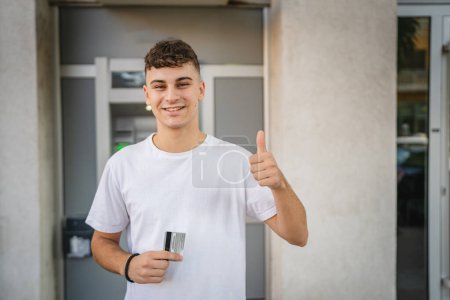Photo for Portrait of teenager man stand in front of bank ATM hold credit card - Royalty Free Image
