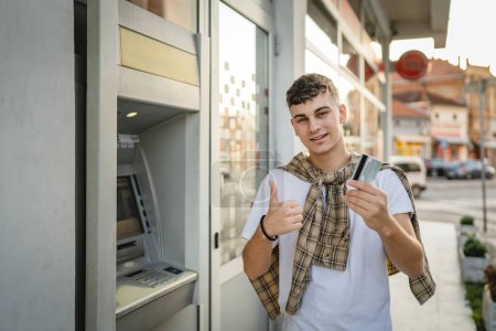 Foto de Retrato de adolescente hombre de pie en frente de banco cajero automático celebrar tarjeta de crédito - Imagen libre de derechos