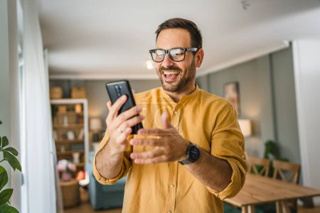 Foto de Un hombre adulto caucásico macho con barba y gafas de pie en casa sonrisa feliz uso de teléfono móvil smartphone para hacer una llamada hablando de espacio de copia de videollamada - Imagen libre de derechos