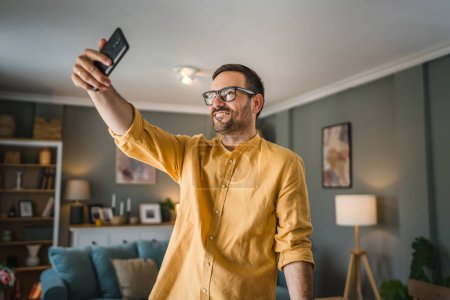 Photo for One man adult caucasian male with beard and eyeglasses stand at home happy smile use mobile phone smartphone to make a call talking video call copy space - Royalty Free Image