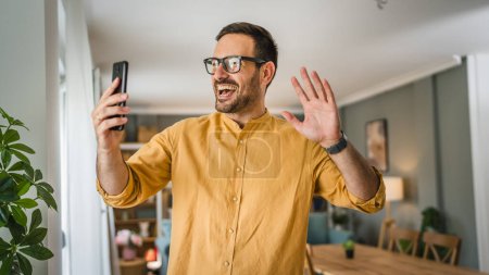 Téléchargez les photos : Un homme adulte caucasien mâle avec barbe et lunettes se tenir à la maison sourire heureux utiliser smartphone téléphone mobile pour faire un appel parler espace de copie d'appel vidéo - en image libre de droit