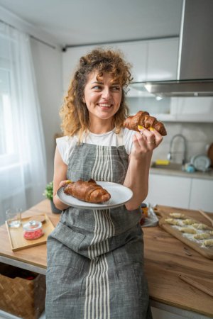 Téléchargez les photos : Une femme caucasienne adulte se tient dans la cuisine avec croissant frais pour le petit déjeuner routine quotidienne du matin - en image libre de droit