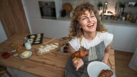 Photo for One adult caucasian woman stand in the kitchen with fresh croissant for breakfast daily morning routine - Royalty Free Image