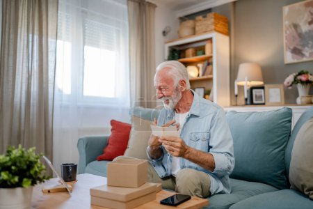 Foto de Un anciano pensionista abuelo con el pelo gris y la barba recibir regalos en la caja abierta tarjeta de lectura sonrisa feliz en frente de la computadora portátil en casa que tiene espacio de copia de videollamada en línea - Imagen libre de derechos