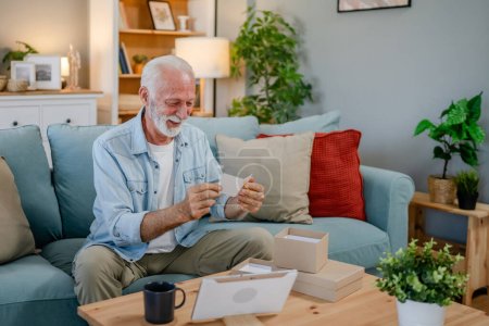 Foto de Un anciano pensionista abuelo con el pelo gris y la barba recibir regalos en la caja abierta tarjeta de lectura sonrisa feliz en frente de la computadora portátil en casa que tiene espacio de copia de videollamada en línea - Imagen libre de derechos