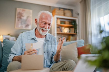 Téléchargez les photos : Un homme âgé grand-père pensionné avec les cheveux gris et la barbe recevoir des cadeaux dans la boîte ouverte carte de lecture heureux sourire devant ordinateur portable à la maison ayant en ligne espace de copie d'appel vidéo - en image libre de droit