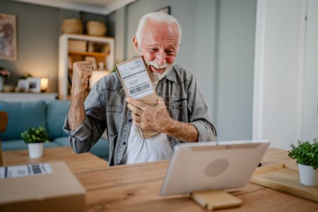 Téléchargez les photos : Un homme caucasien senior à la maison tenir paquet cardbox livré cadeau ou une commande utiliser une tablette numérique en ligne pour vérifier l'état des achats d'expédition ou pour l'espace de copie d'appel vidéo - en image libre de droit