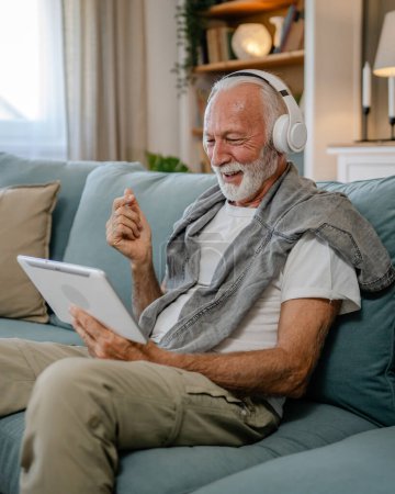 Photo for One senior man old caucasian male pensioner sit at home on the sofa bed use digital tablet to watch movie or have a video call online with headphones on his head gray hair and beard - Royalty Free Image