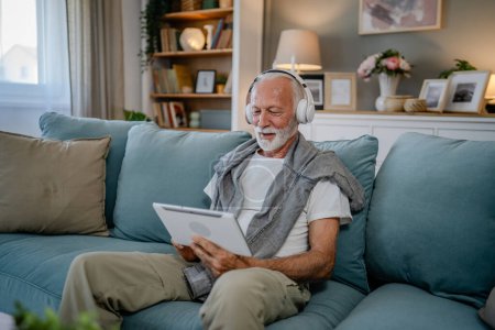 Téléchargez les photos : Un vieil homme âgé retraité caucasien assis à la maison sur le canapé-lit utiliser une tablette numérique pour regarder un film ou avoir un appel vidéo en ligne avec des écouteurs sur sa tête cheveux gris et la barbe - en image libre de droit