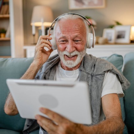 Téléchargez les photos : Un vieil homme âgé retraité caucasien assis à la maison sur le canapé-lit utiliser une tablette numérique pour regarder un film ou avoir un appel vidéo en ligne avec des écouteurs sur sa tête cheveux gris et la barbe - en image libre de droit
