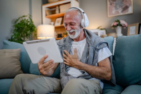 Photo for One senior man old caucasian male pensioner sit at home on the sofa bed use digital tablet to watch movie or have a video call online with headphones on his head gray hair and beard - Royalty Free Image