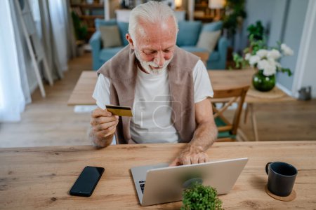Téléchargez les photos : Un homme caucasien senior grand-père masculin assis à la maison utiliser carte de crédit et ordinateur portable achats en ligne utiliser Internet pour l'achat en ligne active senior moderne - en image libre de droit