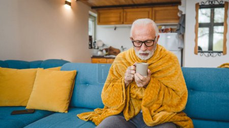 Téléchargez les photos : Un homme âgé caucasien assis sur le canapé-lit à la maison tenir tasse de thé froid commun éternuer malade attrapé froid - en image libre de droit