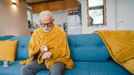 Foto de Un hombre mayor caucásico se sienta en el sofá cama en casa mantenga el termómetro comprobar la temperatura corporal fiebre o resfriado común estornudos enfermos atrapados frío - Imagen libre de derechos