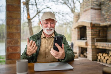 Téléchargez les photos : Un homme homme caucasien senior avec barbe grise et casquette assis en plein air au printemps ou en automne jour heureux sourire utiliser téléphone intelligent mobile pour l'espace de copie d'appel vidéo vraies personnes - en image libre de droit