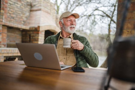 Photo for One senior man sit outdoor in autumn day hold cup of coffee or tea use laptop computer - Royalty Free Image