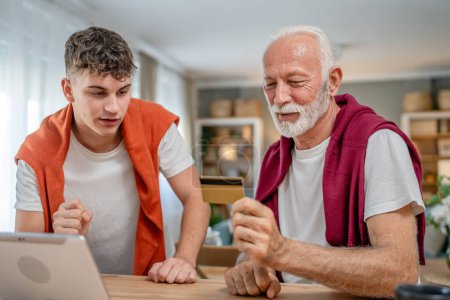 Téléchargez les photos : Un adolescent jeune homme et vieux caucasien mâle grand-père assis à la maison utiliser carte de crédit et ordinateur portable achats en ligne utiliser Internet pour l'achat en ligne active senior moderne - en image libre de droit