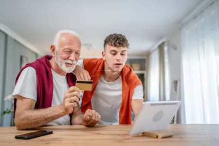 Téléchargez les photos : Un adolescent jeune homme et vieux caucasien mâle grand-père assis à la maison utiliser carte de crédit et ordinateur portable achats en ligne utiliser Internet pour l'achat en ligne active senior moderne - en image libre de droit