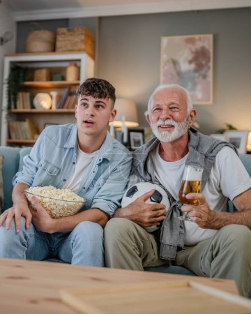 Foto de Caucásico adolescente y hombre mayor abuelo y nieto sentarse en el sofá cama en casa ver fútbol partido de fútbol celebrar palomitas de maíz tazón y vaso de cerveza divertirse familia vinculación hombres - Imagen libre de derechos