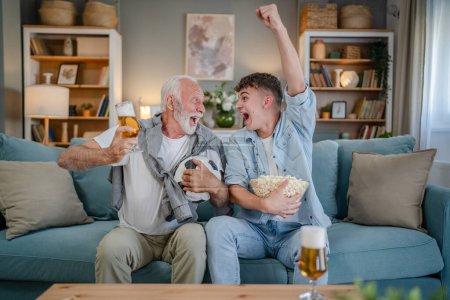 Photo for Caucasian teenager and senior man grandfather and grandson sit on sofa bed at home watch football soccer game hold popcorn bowl and glass of beer have fun family bonding males men - Royalty Free Image