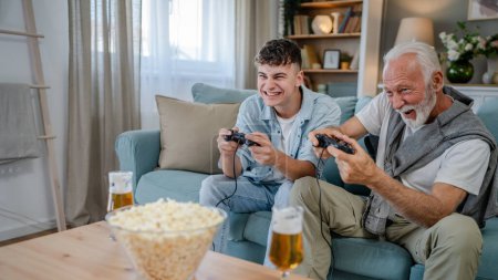 Photo for Caucasian teenager and senior man grandfather and grandson sit on sofa bed at home play console video game hold joystick controller have fun family bonding males men - Royalty Free Image