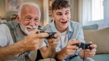 Photo for Caucasian teenager and senior man grandfather and grandson sit on sofa bed at home play console video game hold joystick controller have fun family bonding males men - Royalty Free Image