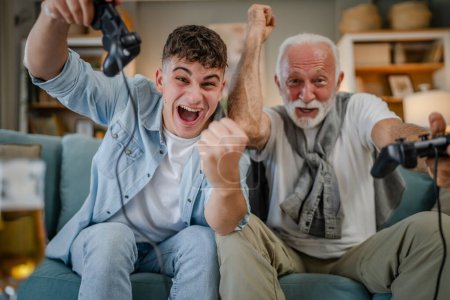 Téléchargez les photos : Adolescent caucasien et senior homme grand-père et petit-fils assis sur le canapé-lit à la maison console de jeu vidéo tenir manette de jeu avoir amusant famille liaison hommes hommes - en image libre de droit