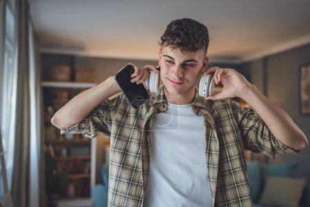 Foto de Retrato de un adolescente parado en casa usar auriculares y smartphone para reproducir música o ver podcast de vídeo en casa - Imagen libre de derechos