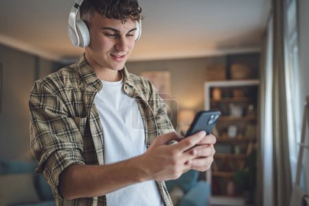 Foto de Retrato de un adolescente parado en casa usar auriculares y smartphone para reproducir música o ver podcast de vídeo en casa - Imagen libre de derechos