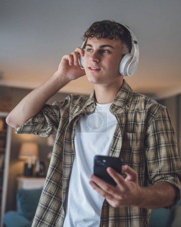 Téléchargez les photos : Portrait de garçon adolescent stand à la maison utilisation casque et smartphone pour jouer de la musique ou regarder podcast vidéo à la maison - en image libre de droit