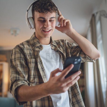 Téléchargez les photos : Portrait de garçon adolescent stand à la maison utilisation casque et smartphone pour jouer de la musique ou regarder podcast vidéo à la maison - en image libre de droit