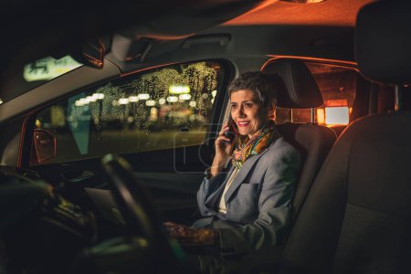 Téléchargez les photos : Une femme âgée mature caucasienne assise dans la voiture travaillant sur ordinateur portable faire un appel téléphonique parler la nuit avec les cheveux gris court espace de copie moderne sourire heureux - en image libre de droit