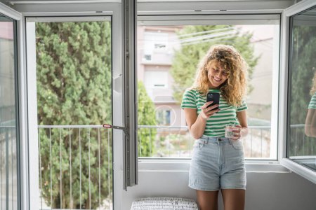 Photo for One woman adult caucasian female stand at the window at home in bedroom use mobile phone hold glass of water send sms texting text messages daily morning routine happy smile copy space - Royalty Free Image