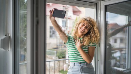 Photo for One woman adult caucasian female stand at window at home in bedroom use mobile phone hold smartphone take selfie photos self portrait or make a video call daily morning routine happy smile copy space - Royalty Free Image