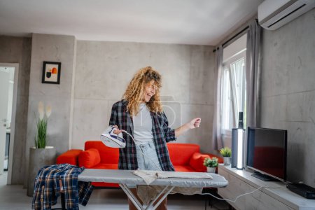 Téléchargez les photos : Femme adulte caucasienne heureuse femme utilisant du fer électrique pour repasser les vêtements à la maison tâches ménagères concept de vraies personnes travail domestique copie espace - en image libre de droit