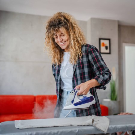 Photo for Caucasian adult woman happy female using electric iron for ironing clothes at home housework household chores concept real people domestic work copy space - Royalty Free Image